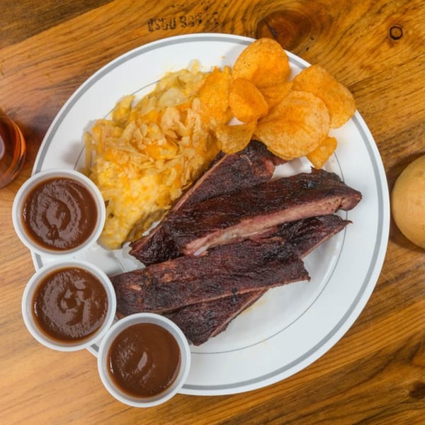 Yazoo BBQ ribs with chips and three small cups of barbecue sauce on a white plate.