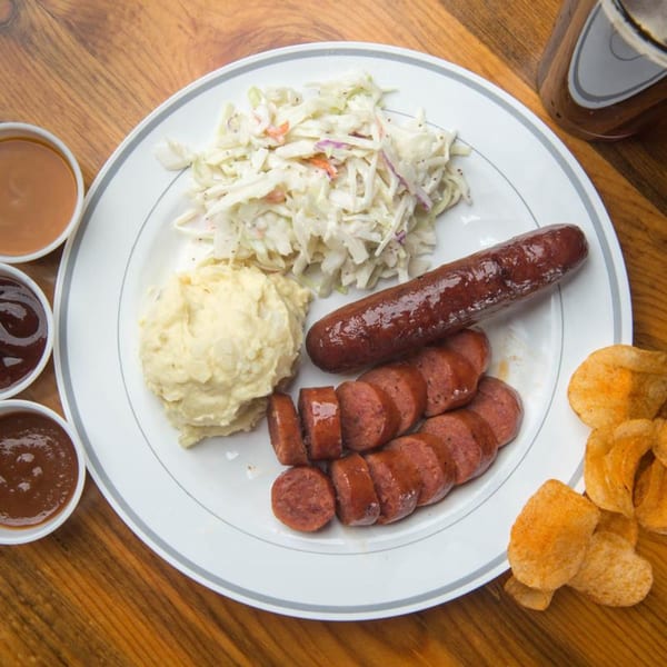 Yazoo BBQ plate with whole and sliced sausage, mashed potatoes, coleslaw, chips, and sauces.