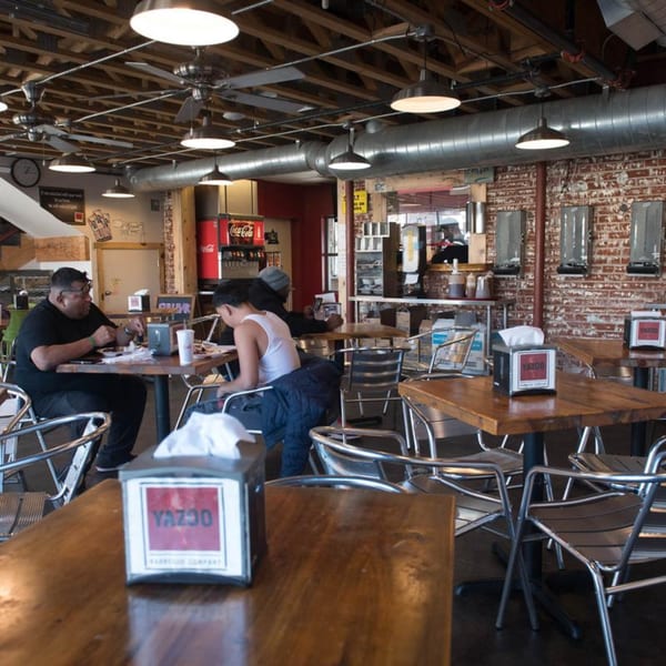 Interior of Yazoo BBQ restaurant with exposed brick, wooden tables, metal chairs, and customers dining.