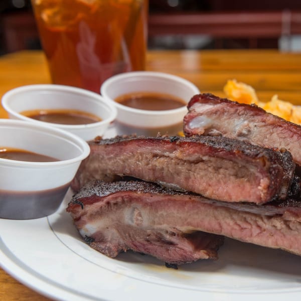 Close-up of Yazoo BBQ ribs with three cups of sauce and a drink in the background.