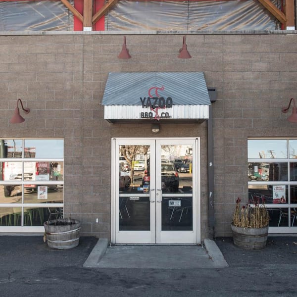 Exterior of Yazoo BBQ Co. restaurant with a gray brick facade, glass doors, and two large windows.