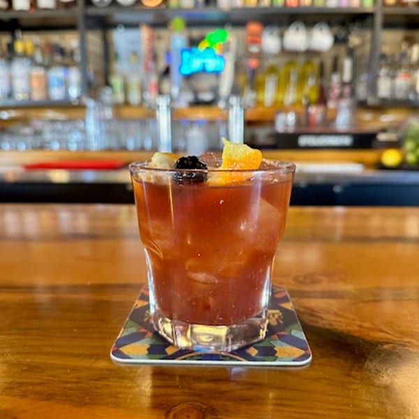 A Yazoo BBQ cocktail with orange and cherry garnish sits on a coaster at a bar.