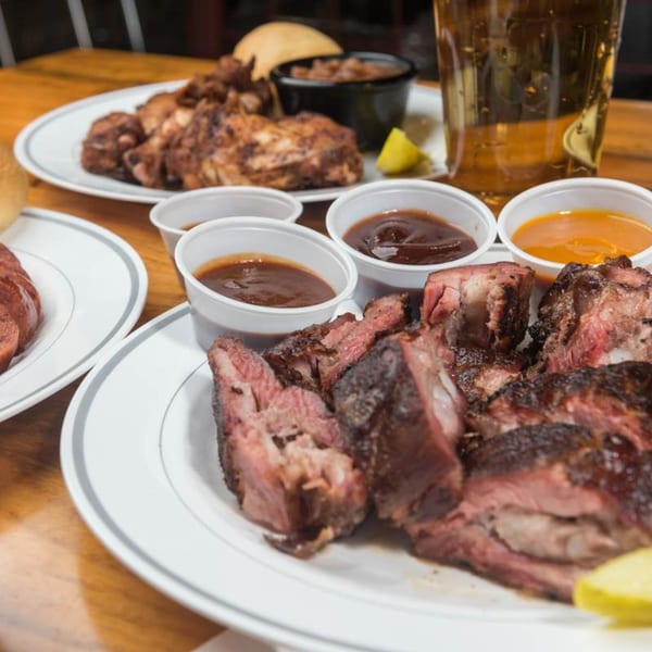 Plates of Yazoo BBQ ribs, chicken, baked beans, and sauces on a wooden table with a beer.