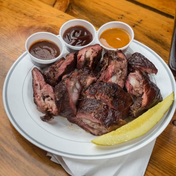 Yazoo BBQ ribs with three sauces and a pickle spear on a white plate.